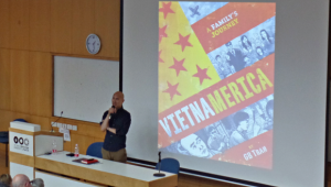 GB Tran giving a lecture and the cover page and title of his book behind him on the screen